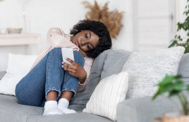 Loneliness. Depressed Black Lady Sitting On Couch With Smartphone, Loking At Screen Loneliness. Depressed Black Lady Sitting On Couch With Smartphone, Loking At Device Screen, Feeling Upset And Bored At Home, Having No Friends, Heartbroken Woman Waiting For Message From Boyfriend symbiotic relationship stock pictures, royalty-free photos & images