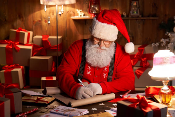 feliz viejo y barbudo santa claus usando sombrero, gafas, escribiendo en la lista de deseos, trabajando en la víspera de navidad sentado en la acogedora mesa del taller casero tarde con regalos, árbol y velas preparándose para las vacaciones. - santa hat christmas hat headwear fotografías e imágenes de stock