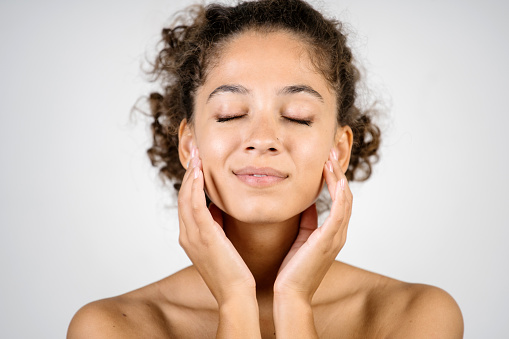 Moisturizing and pampering concept. Portrait of smiling adult african american woman touching clear face skin with hands, standing in spa salon isolated on white background with copy space