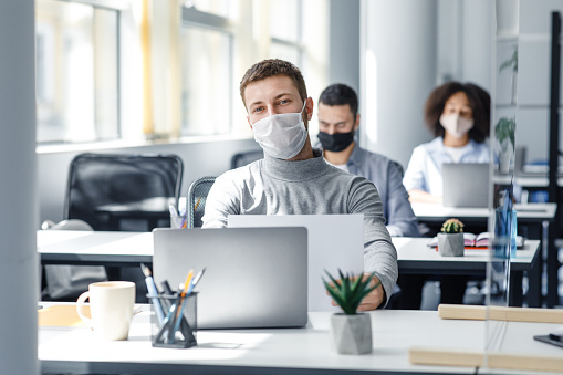 Business and coronavirus epidemic after lockdown. Millennial male worker in protective mask works with documents and laptop at workplace with protective glass and searching information