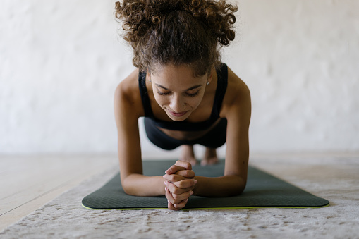 Smiling young afro american sportswoman training at home, making sport exercise, standing in plank pose on yoga mat against blurred copy space background