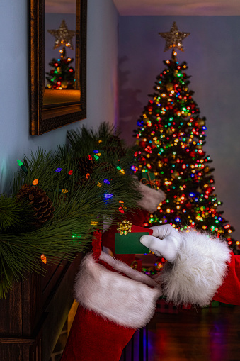 Santa Claus’s gloved hand placing 2 gift cards into a Christmas stocking hanging on a fireplace mantel with a Christmas tree in the background surrounded with wrapped presents on Christmas eve.