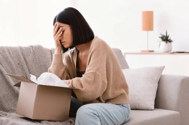 Wrong Item Concept. Sad unpset asian lady sitting on couch and unpacking cardboard box, received parcel with damaged staff, covering face in disappointment, feeling dissatisfied with bad purchase