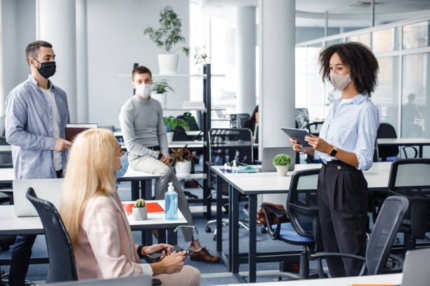 reunião corporativa e trabalho em grupo na empresa. mulher afro-americana gerente em máscara protetora segurando tablet, falando com trabalhadores mantendo distância social - distanced - fotografias e filmes do acervo