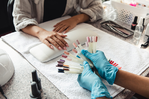 Choose of trendy color in beauty studio after reopening. African american woman in protective gloves showing samples of nail polish to client on table with nail equipment during covid-19, cropped