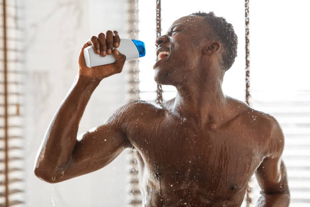 homme africain positif chantant prenant la douche retenant la bouteille de shampooing à l’intérieur - men naked water male photos et images de collection