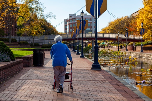 Frederick, MD, 10/13/2020: An elderly caucasian woman wearing blue sweater, track pants and sneakers is slowly walking in parks with a rollator walker. It is a sunny autumn day in Frederick, Maryland with river on one side