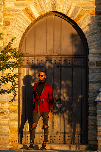 Frederick, MD, USA 10/14/2020:  African American male model wearing fashionable red jacket, sun glasses and ripped jeans poses at a historic place with arched door and stone wall behind him at sunset