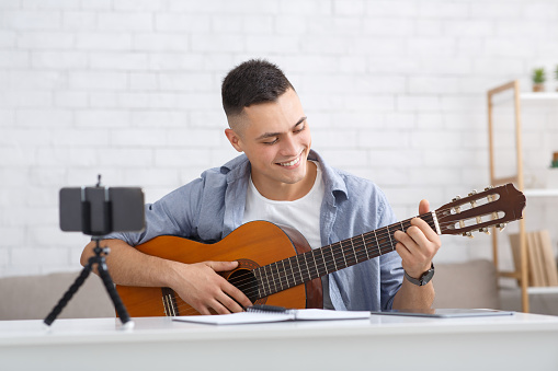 Young student learning to play guitar during self-isolating at home or online tutor. Smiling guy looks at guitar and makes video lesson for followers in living room during virus outbreak, free space