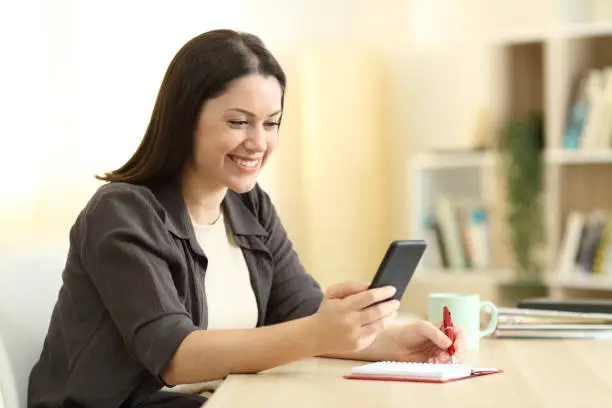 Photo of Happy woman writing in agenda and checking phone