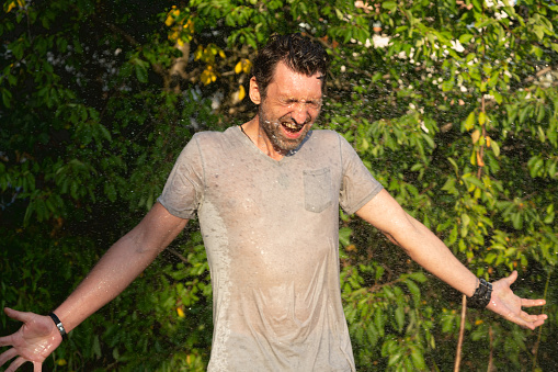 Midadult man standing in garden with outstretched arms while there is water splashed at him