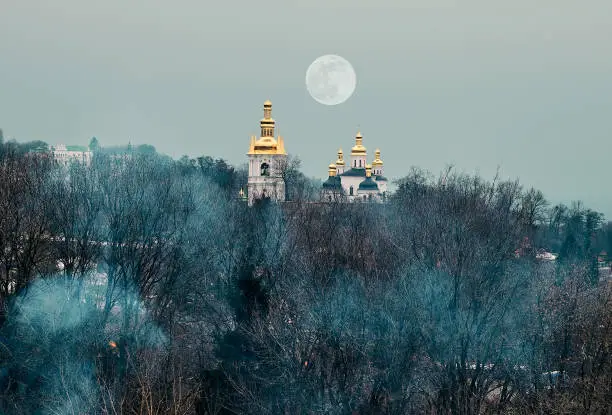 Photo of Blue smoke envelops trees without leaves. In the distance you can see the temple of God. Golden and green domes with church crosses. Cloudy autumn day. The moon hung over the grove.