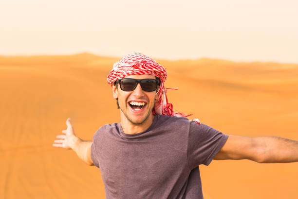 Happy young male tourist enjoying a trip in the Arabian desert. Cheerful and excited caucasian young man standing with arms open and smiling at the camera against the golden desert sand dunes in the background. He is wearing the local headwear and sunglasses. arabian sunglasses stock pictures, royalty-free photos & images