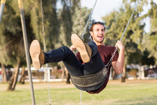 Happy and cheerful young caucasian male adult enjoying riding a swing at the park's playground during a sunny summer day.