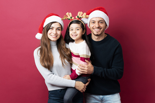 Hispanic family of three hugging each other standing against a red background on Christmas