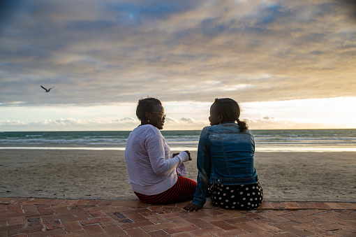 An African Ethnicity Mother sitting talking to her teenage daughter at the ocean at sunset with a seagull flying by