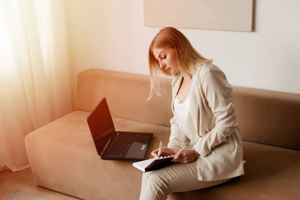 istock Woman is using a laptop while sitting on the couch holding a diary and a pen planning day timetable, businesswoman concept, working from home 1281692089