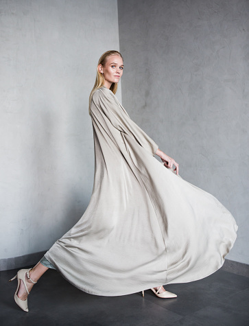 Gorgeous young blonde woman walking and posing in a studio while wearing a luxury abaya made of a long grey fabric.
