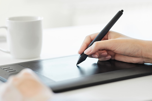 Close up of a hand holding a pen and drawing on a tablet surface retouching, drawing or making a graphic design