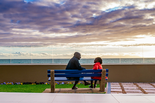 A Nigerian African Ethnicity Father and his daughter rear view sitting on a bench bonding talking sharing with a sunset over the ocean in front of them.