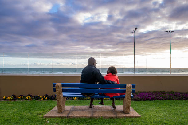 A Nigerian African Ethnicity Father and his daughter rear view sitting on a bench bonding talking sharing with a sunset over the ocean in front of them.