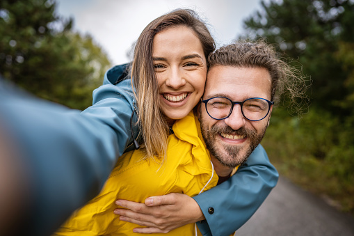 Photo of young love couple having fun while piggybacking in nature and taking selfies with smart phone.