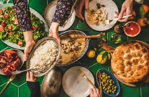 Family having Turkish dinner. Flat-lay of people passing rice pilav over green table with lamb in yogurt sauce, fresh arugula and strawberry salad and flatbread, top view. Ramazan iftar supper