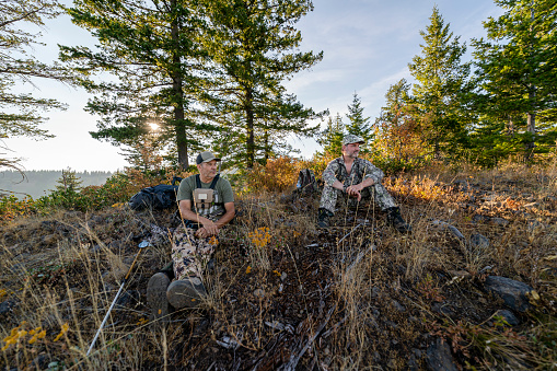 Two middle aged men wearing camouflage clothing sit on a mountain peak in Washington state while hunting elk with a crossbow. It is a warm and sunny Autumn afternoon.