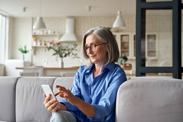 istock Relaxed mature old 60s woman, older middle aged female customer holding smartphone using mobile app, texting message, search ecommerce offers on cell phone technology device sitting on couch at home. 1281725865