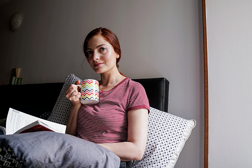 Latin woman of average age 25 years old with red hair dressed in pajamas is sitting on her bed has a cup of coffee and enjoys the moment alone as daily rituals