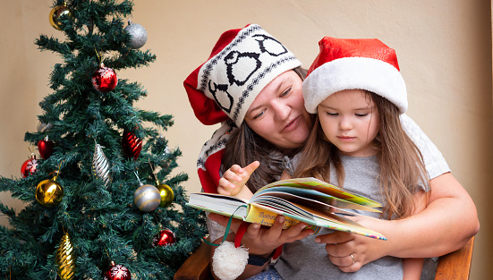 mother and daughter are reading a book sitting in a chair near the christmas tree in santa hat