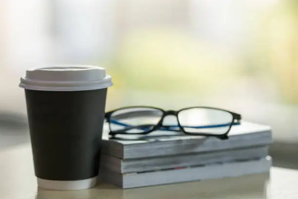 Closeup of takeaway papercup of hot coffee with stack of books and reading glasses on wooden table.