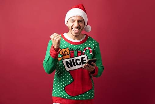 Handsome Latin man celebrating and looking excited about Christmas while using a smartphone in a studio