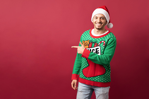 Cheerful Latin man wearing ugly sweater and pointing towards copy space in a studio against a red background