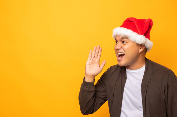 istock Asian man wearing christmas hat and shouting with hand into blank space for text advertise. Merry christmas and happy new years people with isolated yellow background. 1281741831