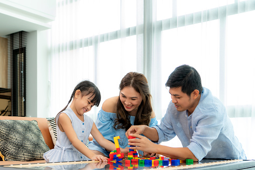 Asian family spends time in playroom with father, mother and daughter with toys on room background build out of plastic blocks in living room at home