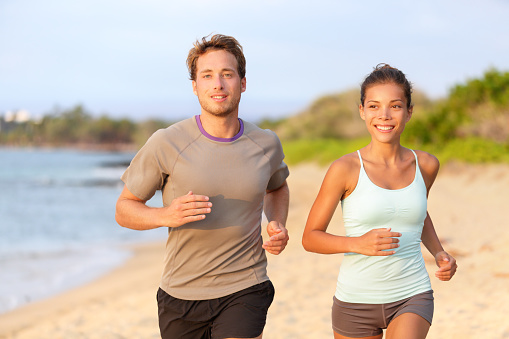 Fitness couple jogging outside on beach smiling in summer sunset. Young mixed race asian female model running on beach with handsome caucasian male.