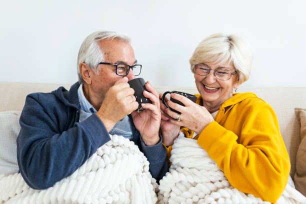 istock Senior couple feeling cozy and warm, sitting covered with blanket on the sofa at home. Elderly couple drinking tea 1281745568
