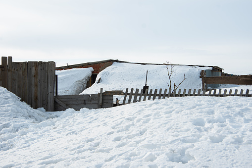 Old village house, old wooden fence covered with snow during a blizzard. The house is covered with snow after a storm or heavy snowfall