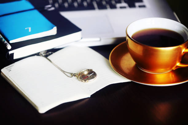 Large golden vintage tea cup with black coffee and open notebook with gold compass jewelry. Laptop and colourful notebooks as backdrops. Coffee break for planning and dreaming time.