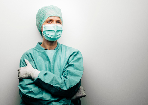 Portrait of healthcare worker wearing protective workwear during coronavirus epidemic. Mature female surgeon is standing with arms crossed. She is against white background.
