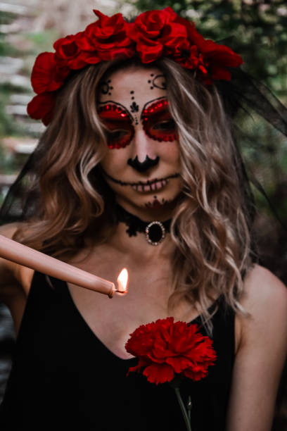 istock Happy Halloween concept. Scared horrible female in special outfit, wears white clay skull, professional sugar skull makeup to look spooky, wears black clothes playing with candle 1281754375