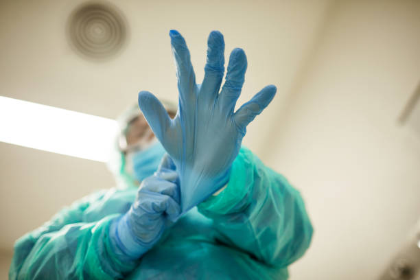 Male Healthcare Worker Putting on Surgical Gloves Low angle personal perspective of male nurse in gown, mask, and eyewear putting on gloves before surgery. frontline worker mask stock pictures, royalty-free photos & images