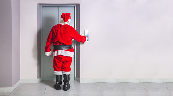 Man disguised as Santa Claus calls the elevator in a building at Christmas in the corridor of a building with a wall with space for text