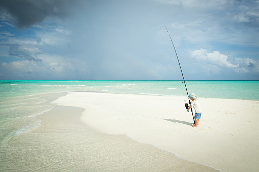 A child fishes on a sandbank in the tropics by himself with a long  fishing rod