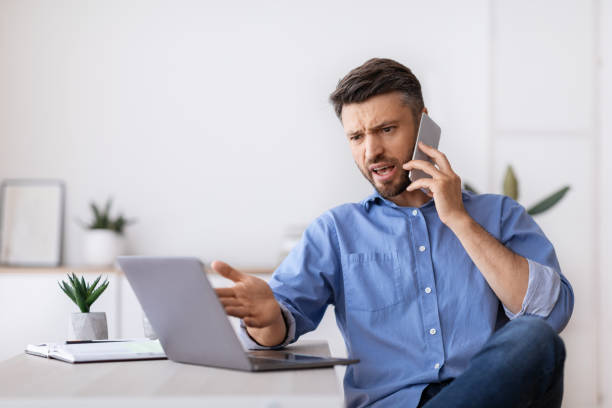 istock Angry businessman talking on cellphone and looking at laptop screen in office 1281768450