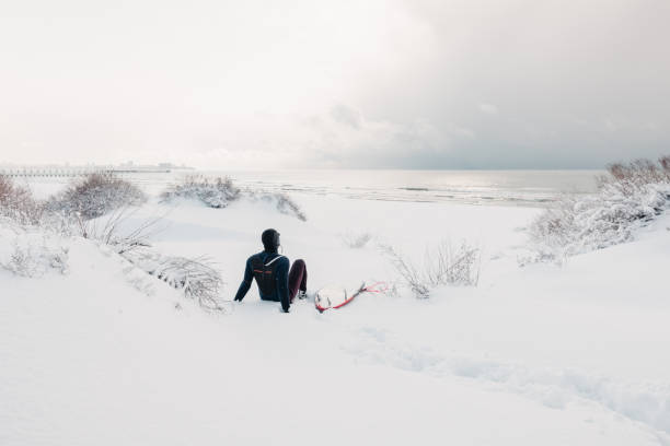 istock Cold winter and surfer sit on snowy beach with surfboard and see at waves. Winter day with surfer in wetsuit. 1281769521