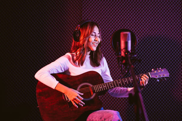 istock Pretty Asian female singer recording songs by using a studio microphone and pop shield on mic while playing guitar and wearing headphones in the studio. Performance and show in the music business. 1281772111