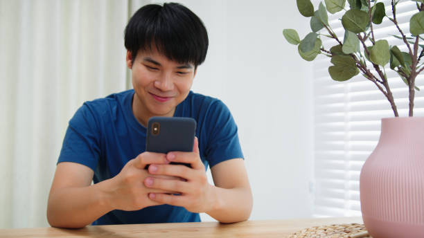 istock Young Asian Man using mobile phone enjoying searching, surfing on the internet on a phone. Happy Man looking at smartphone texting messages, chatting and Smile Sitting at home 1281780289