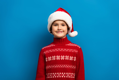 happy boy in santa hat and red sweater looking at camera on blue